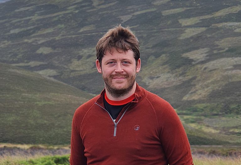 Photo of George Deeks in a red sweatshirt against a background of hills
